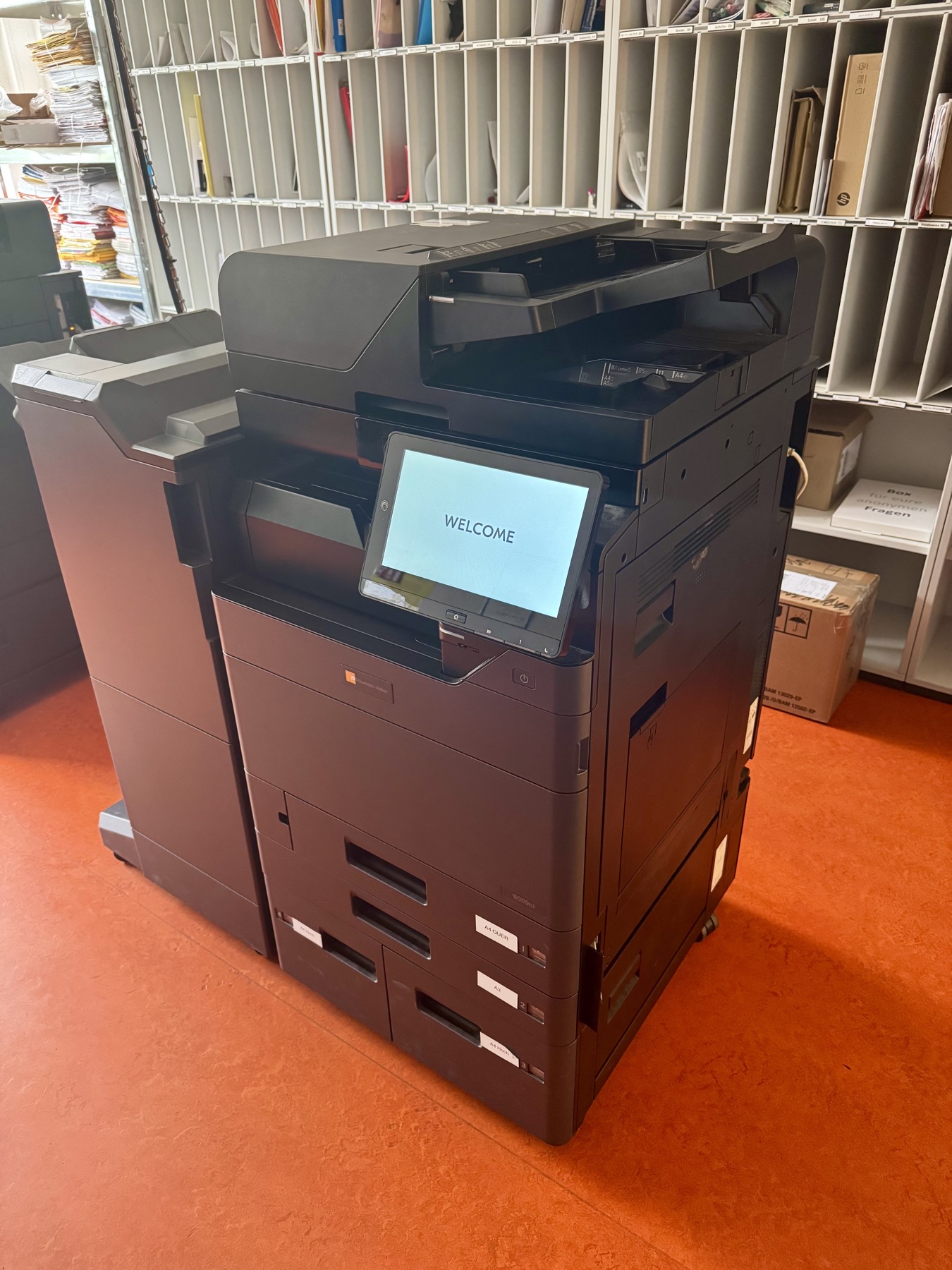 Triumph-Adler 6009ci im Lehrerzimmer, daneben ein Finisher, der Touchscreen zeigt „Welcome“
