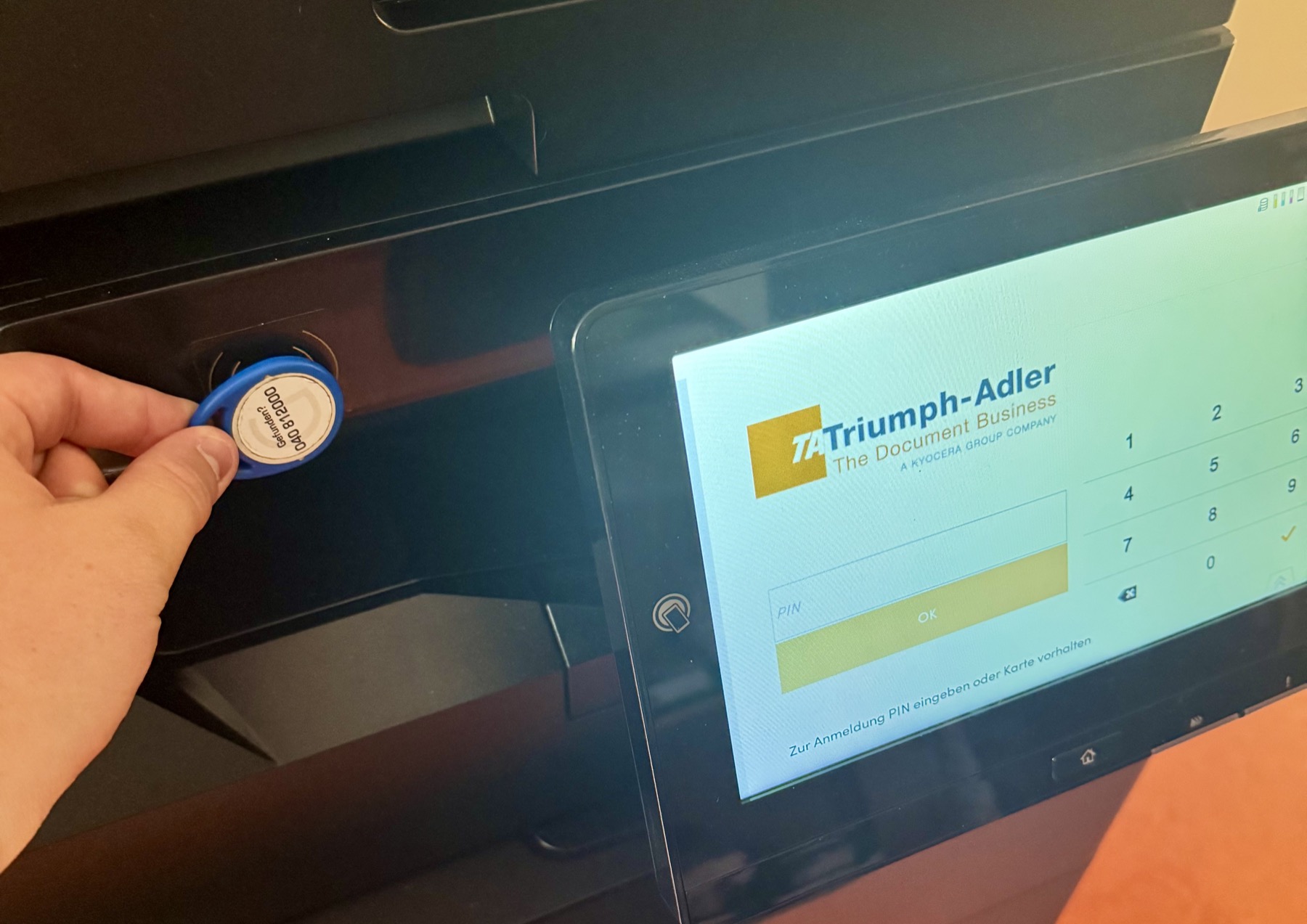Blauer Druckerschlüssel wird an das Lesegerät links neben dem Touchscreen gehalten, auf dem Bildschirm die Anmeldemaske
